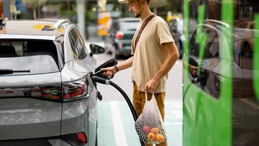 Man with groceries waiting for electric car to be charged on public charging station near a shopping area